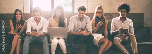 Group of confident young people using different gadgets while sitting close to each other in a row