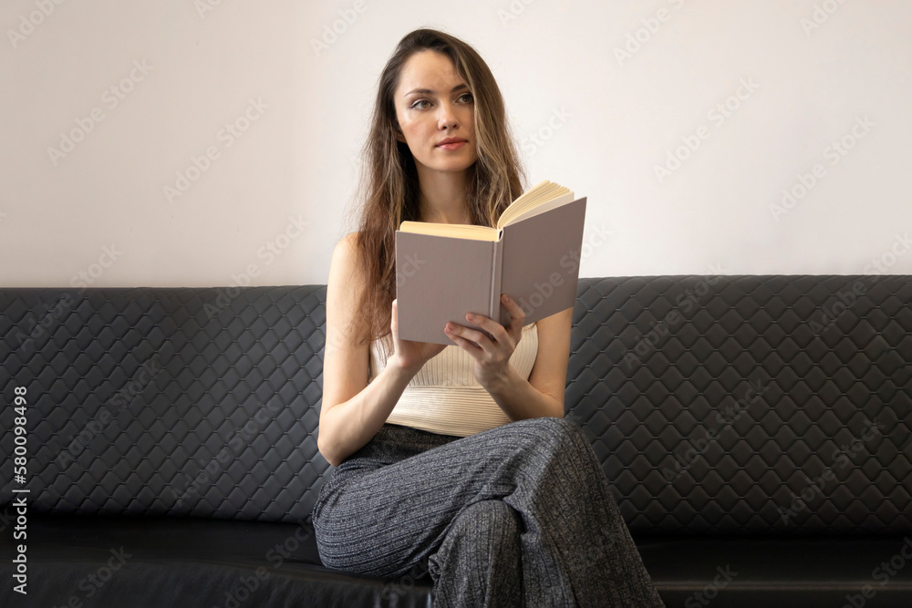 Beautiful, brooding young woman sits on the couch, holds an open book, and looks away Beautiful, brooding young woman sits on the couch, holds an open book, and looks away
