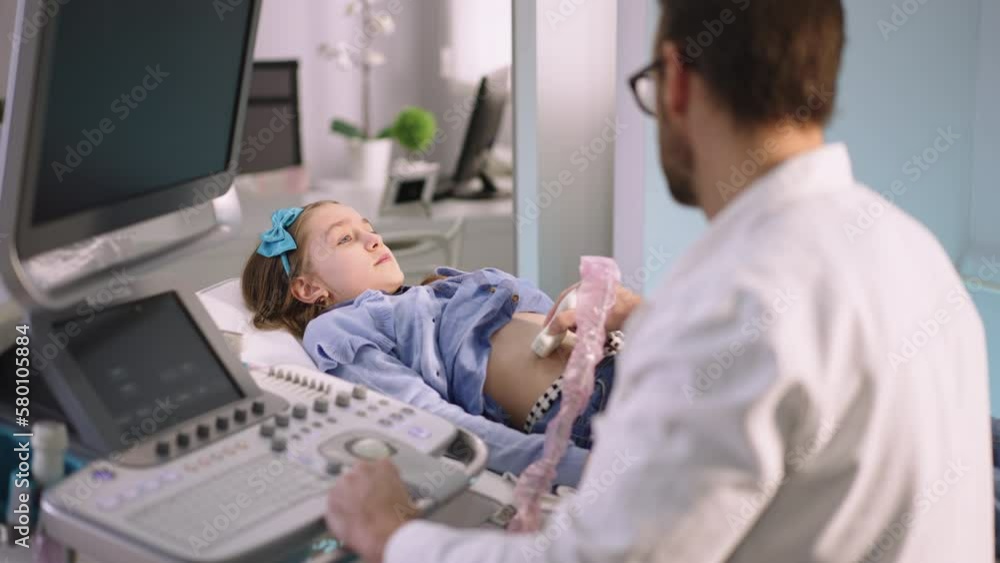 Young bearded doctor is examining little girl's belly by using an ultrasound equipment in clinic Young bearded doctor is examining little girl's belly by using an ultrasound equipment in clinic