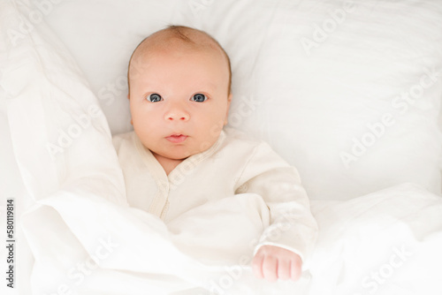 Cute baby wake up in bed on white duvet with surprised face top view closeup. Childhood. Good morning.