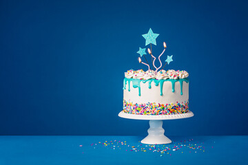 White birthday drip cake with teal ganache, star toppers and fun candles over dark blue background