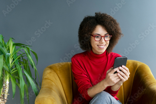 Smiling African American woman holding smartphone watching videos sitting on armchair at home. Happy modern female using mobile app shopping online, ordering food, reading text message, check email © Maria Vitkovska