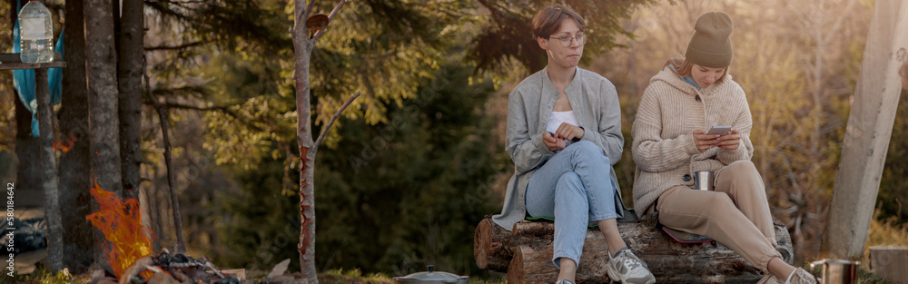 Two young Caucasian women sitting at fire in forest and resting after hiking. Females tourists. Two young Caucasian women sitting at fire in forest and resting after hiking. Females tourists.