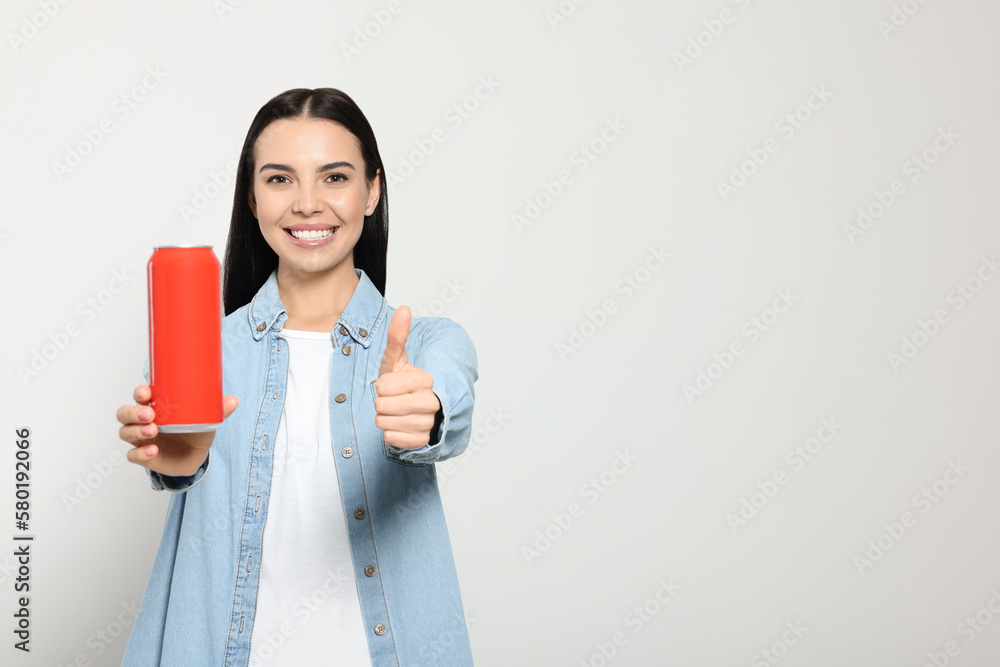 Beautiful happy woman holding red beverage can and showing thumbs up on light grey background. Space for text Beautiful happy woman holding red beverage can and showing thumbs up on light grey background. Space for text
