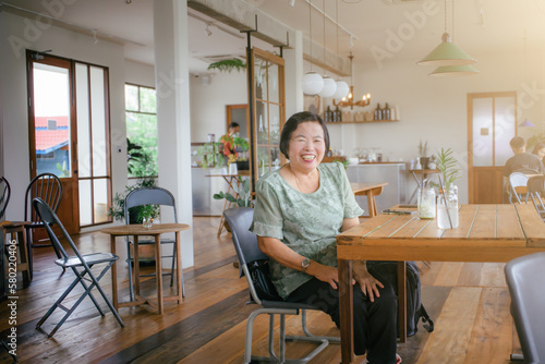 An elderly Asian woman in a traditional green shirt sitting on a chair smiling happily in a cozy cafe.