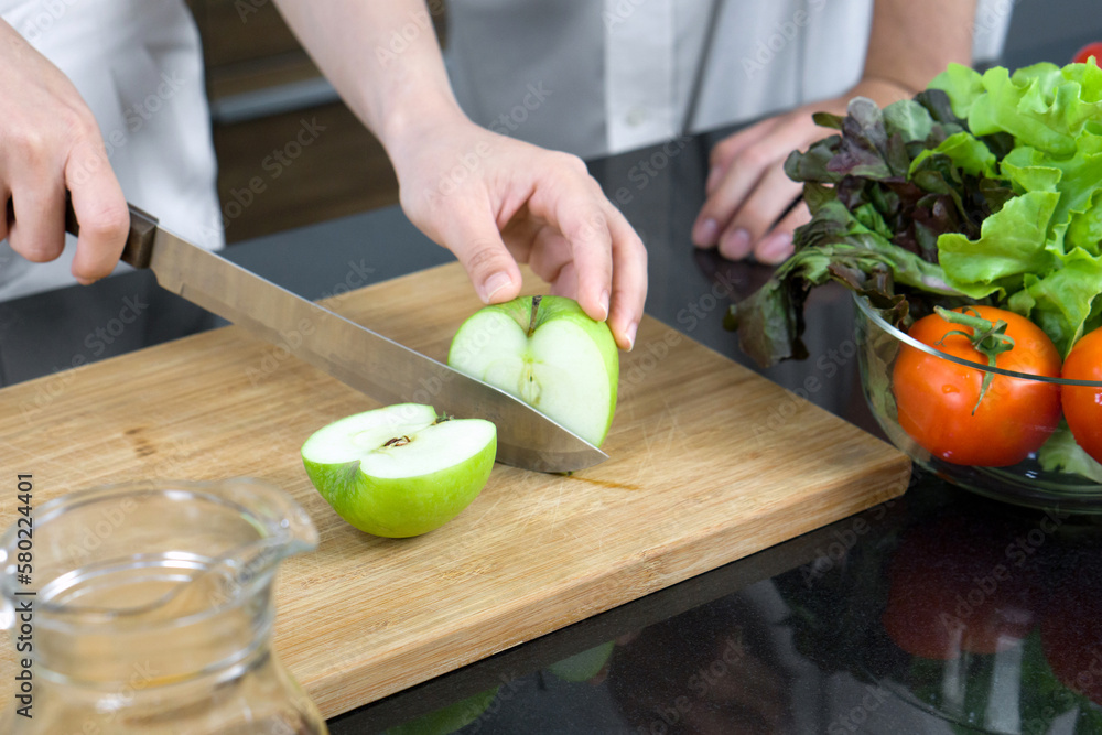 Closeup hand holding knife cutting green apple on a wooden chop board. A glass bowl with a variety of vegetables is placed on the kitchen counter. Closeup hand holding knife cutting green apple on a wooden chop board. A glass bowl with a variety of vegetables is placed on the kitchen counter.