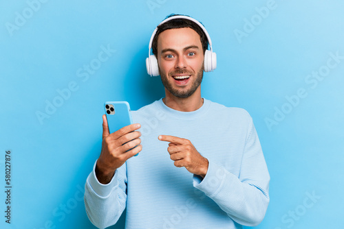 Excited man listening to the music and pointing finger to the smartphone with happy and smiling face isolated over blue background