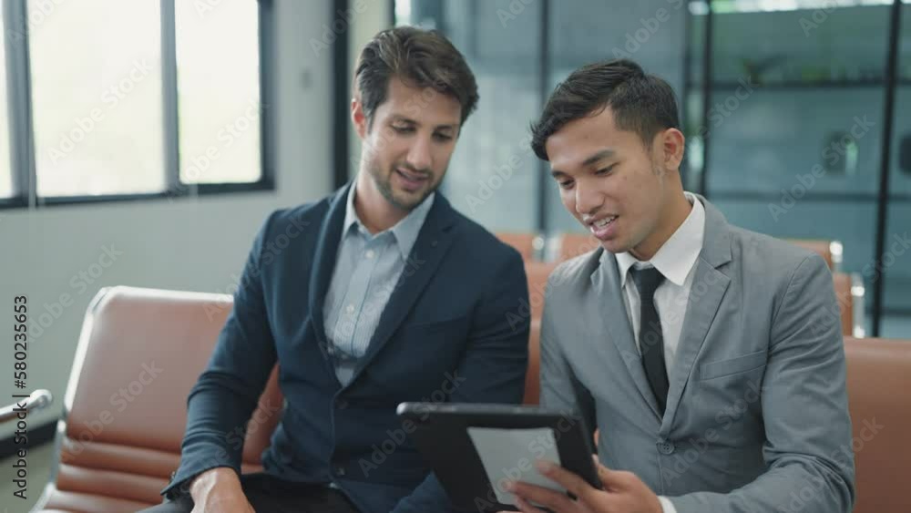 Two businessman wearing suit sitting and talking for work on tablet waiting for flight at airport terminal Two businessman wearing suit sitting and talking for work on tablet waiting for flight at airport terminal