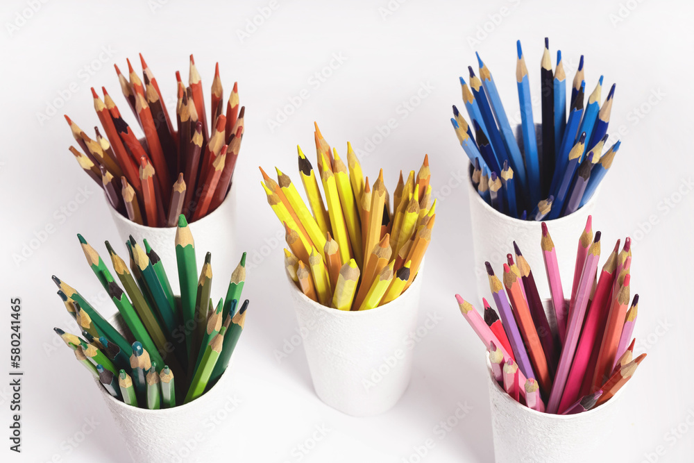 colored pencils in white glasses on a white background. Top view photo of stacks of pencils by color red, yellow, blue-green horizon, pink and purple