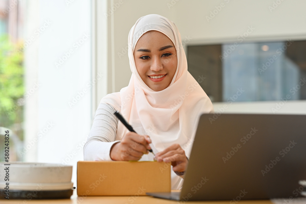 Beautiful Muslim female business owner writing address on cardboard boxes for delivery to customer at e-commerce store. Beautiful Muslim female business owner writing address on cardboard boxes for delivery to customer at e-commerce store.