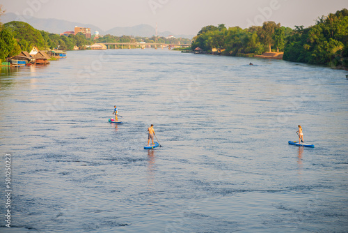 People playing stand-up paddleboarding (SUP) in the Kwai River near the bridge over the Kwai photo