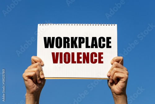 Workplace violence text on notebook paper held by 2 hands with isolated blue sky background. This message can be used as business concept about workplace violence.
