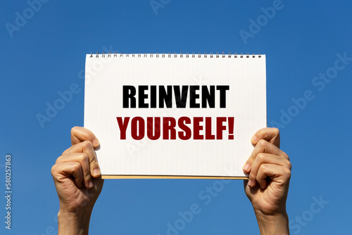 Reinvent yourself text on notebook paper held by 2 hands with isolated blue sky background. This message can be used as business concept to encourage audience reinventing yourself. photo