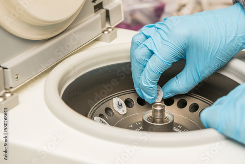 A part of an arm with labcoat and with blue nitril gloves putting a small plastic test tube in a micro centrifuge.