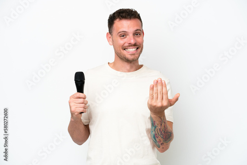 Young caucasian singer man picking up a microphone isolated on white background inviting to come with hand. Happy that you came