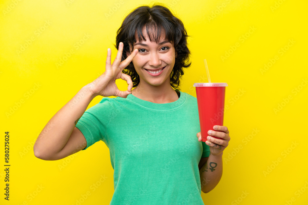 Young Argentinian woman holding a soda isolated on yellow background showing ok sign with fingers Young Argentinian woman holding a soda isolated on yellow background showing ok sign with fingers