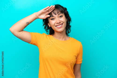 Young Argentinian woman isolated on blue background saluting with hand with happy expression
