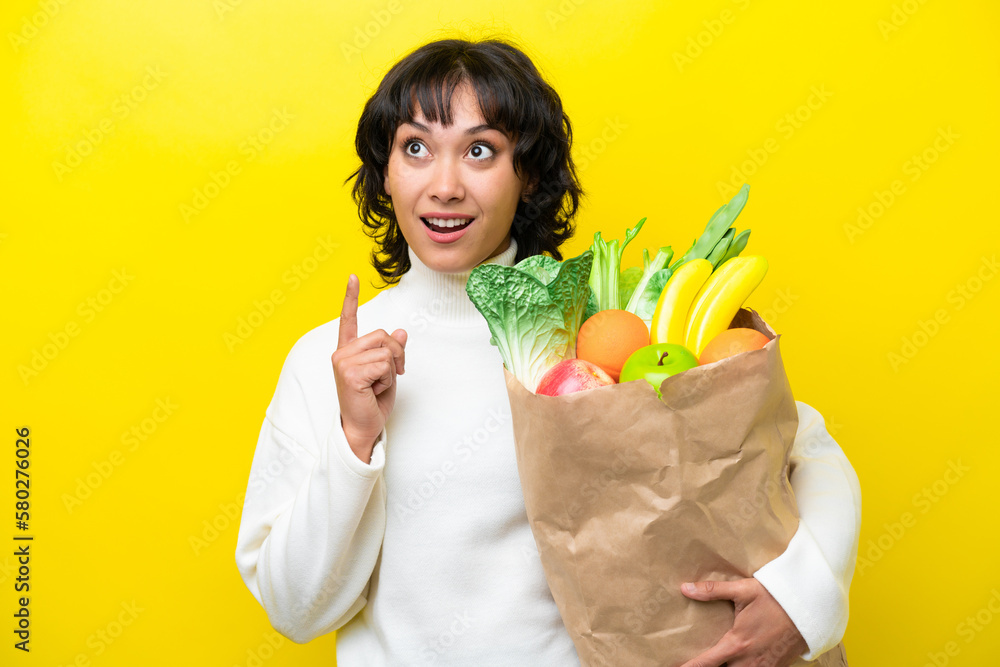 Young Argentinian woman holding a grocery shopping bag isolated on yellow background thinking an idea pointing the finger up Young Argentinian woman holding a grocery shopping bag isolated on yellow background thinking an idea pointing the finger up
