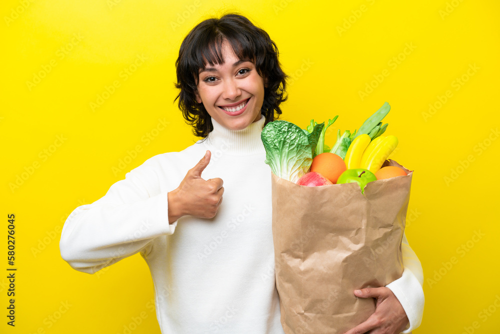 Young Argentinian woman holding a grocery shopping bag isolated on yellow background giving a thumbs up gesture Young Argentinian woman holding a grocery shopping bag isolated on yellow background giving a thumbs up gesture