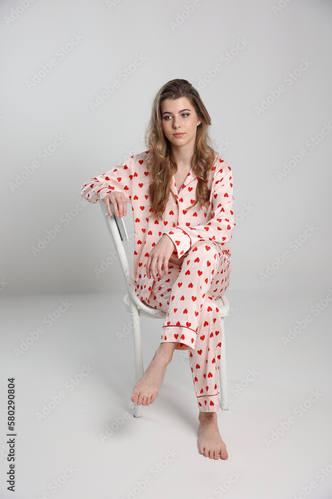 beautiful blonde girl sits on a chair on a white background in pajamas. pajamas with hearts. Valentine's Day. studio photo shoot in satin pajamas with a heart print. clothes for sleep and home