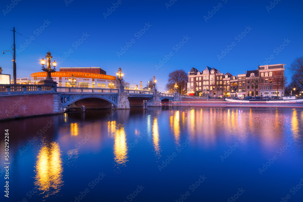 Obraz premium Blue bridge, Amsterdam, Netherlands. Blauwbrug. Evening cityscape. Blue sky and city lights. Dutch canals. Reflections on the surface of the water. Photography for design and wallpaper.
