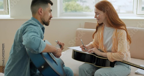 awesome happy smiling girl holding pen, sheet of paper listening her boyfriend who is sitting with guitar, family writing music at home during coronovirus, covid-19 happiness slow motion conversation photo