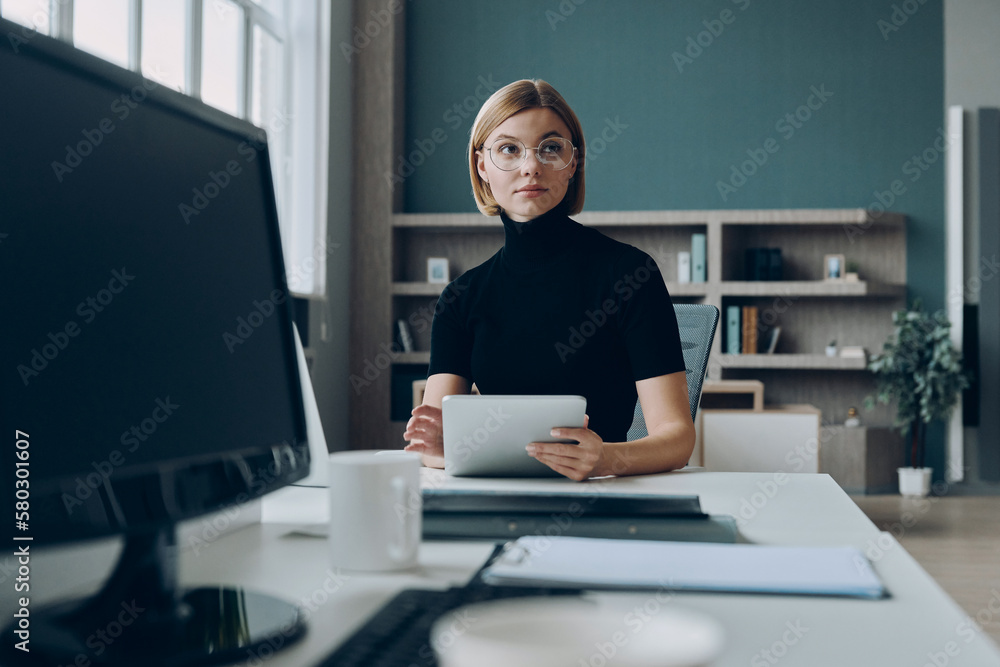 Confident businesswoman holding digital tablet while sitting at her working place in office Confident businesswoman holding digital tablet while sitting at her working place in office
