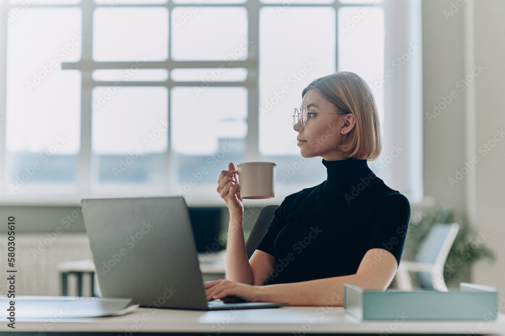 Beautiful young businesswoman holding coffee cup while sitting at her working place in office Beautiful young businesswoman holding coffee cup while sitting at her working place in office