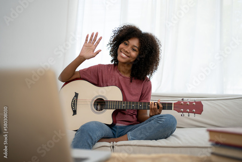 Focused African girl playing acoustic guitar and watching online course on laptop while practicing at home. Online training, online classes.