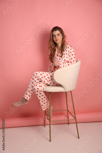 beautiful woman in pajamas with a heart print on a pink background is sitting on a chair. Pink pajamas with red hearts. clothes for sleep and home. Valentine's Day.