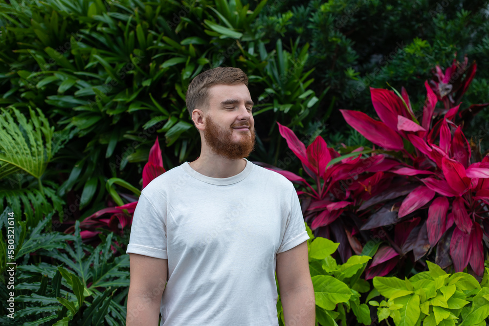 Portrait of happy young bearded man with beard breathing deep, inhale fresh air on natural background, green wall with leaves, flower in tropical country in garden. Eco, ecological concept. Portrait of happy young bearded man with beard breathing deep, inhale fresh air on natural background, green wall with leaves, flower in tropical country in garden. Eco, ecological concept.