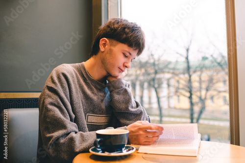 Teenage blond boy 18-19 year old reading book and drinking coffee in cafe alone.