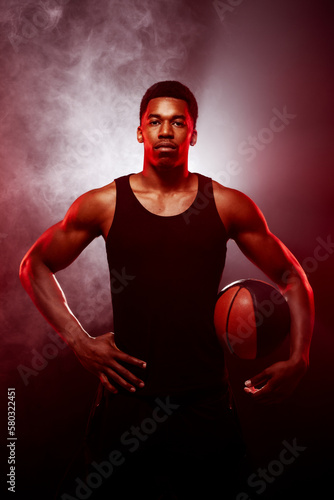 Basketball player side lit with red color holding a ball against smoke background. Serious concentrated african american man..