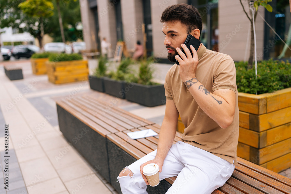 Handsome tattooed young man talking on smartphone holding in hand takeaway coffee sitting on bench on city street. Bearded male having conversation on phone outdoors. Concept of urban lifestyle. Handsome tattooed young man talking on smartphone holding in hand takeaway coffee sitting on bench on city street. Bearded male having conversation on phone outdoors. Concept of urban lifestyle.