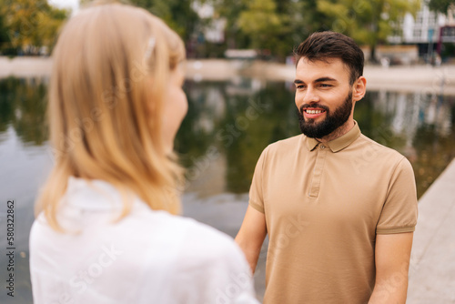 Cheerful bearded man standing holding hands with girlfriend outdoors by beautiful lake at city park, talking looking at each other. Happy bearded man and blonde woman walking spending time together. © dikushin