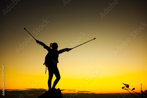 Silhouette of a hiker girl on a rock pedestal with hands up. Beautiful orange sunset.