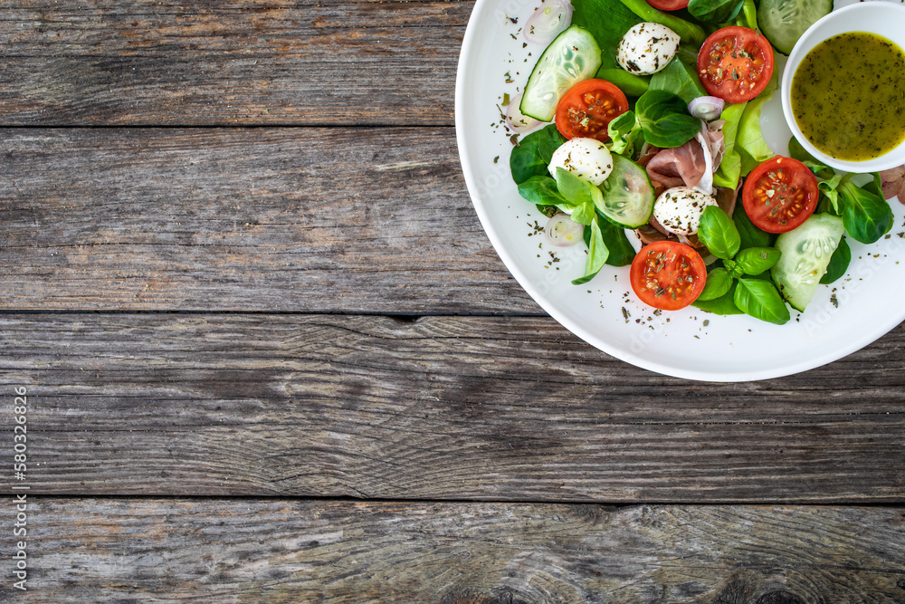 Fresh salad - prosciutto di Parma, mozzarella, cucumber, tomatoes and leafy vegetables on wooden table
