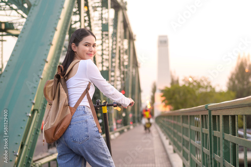 Portrait of young beautiful woman with an electric scooter over bridge in modern city background