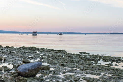 Morning atmosphere at Lake Garda. The water level is historically low, so rocks and stones on the shore stand out. In the background the water, boats, buoys and the shore in the morning light
