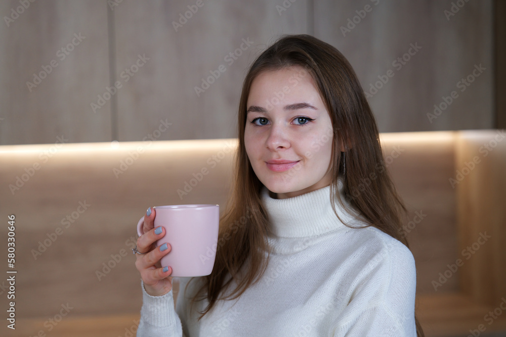 Portrait of happy beautiful girl, young woman with a cup of tea or coffee standing at home at kitchen, smiling in sweater at winter cold day Portrait of happy beautiful girl, young woman with a cup of tea or coffee standing at home at kitchen, smiling in sweater at winter cold day