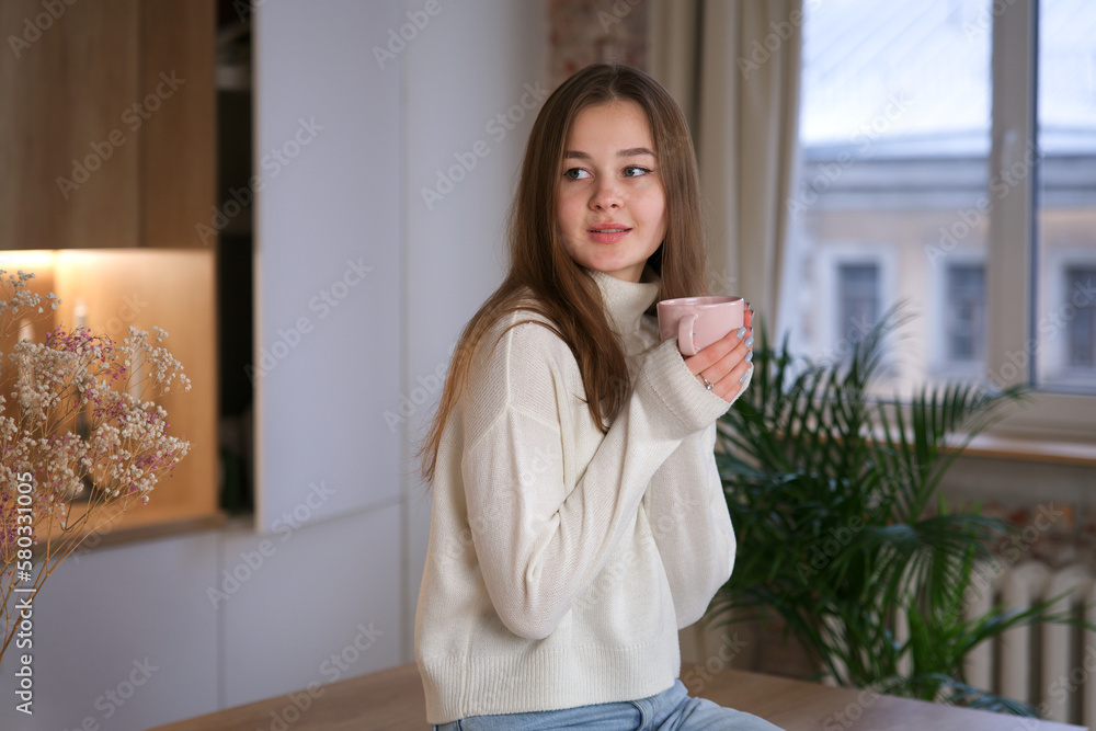 Young beautiful happy woman enjoy with a cup of tea or coffee at kitchen at home in sweater, smile, look at camera Young beautiful happy woman enjoy with a cup of tea or coffee at kitchen at home in sweater, smile, look at camera