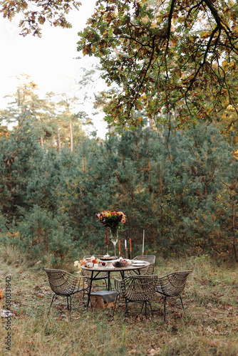 Cozy autumn picnic in the park. Festive setting table decorated wildflowers in vase, candles, food, hot drinks, maples leaves and wicker chairs outdoors. Straw baskets and pumpkins on the grass