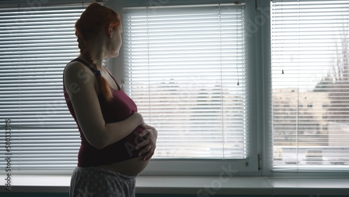 A young pregnant woman in the hospital in anticipation of childbirth looks out the window and strokes her stomach. Experiences of the mother.