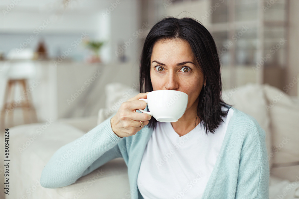 Image of shocked funny female of 50 in casual clothes drinking tea from white cup looking at camera with surprised big round eyes, sitting on couch in living-room, amazed with good news Image of shocked funny female of 50 in casual clothes drinking tea from white cup looking at camera with surprised big round eyes, sitting on couch in living-room, amazed with good news
