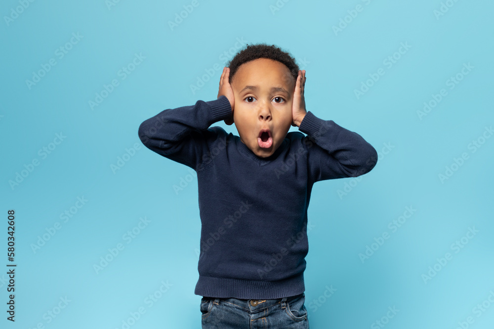 Emotional black boy with his mouth wide open, screaming and closing ears, from what he hear, blue background Emotional black boy with his mouth wide open, screaming and closing ears, from what he hear, blue background