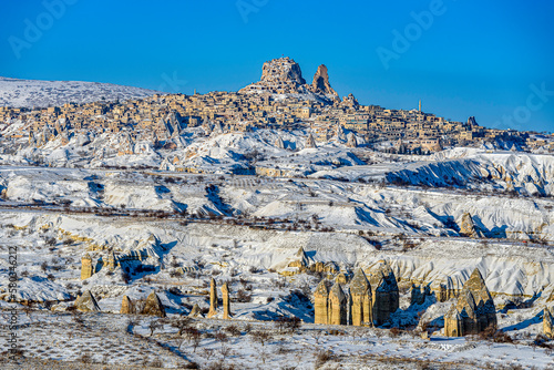 view of snow covered Cappadocia towards Uchisar. The eroded soil is covered with snow. The sun is shining and the sky is blue with fluffy white clouds