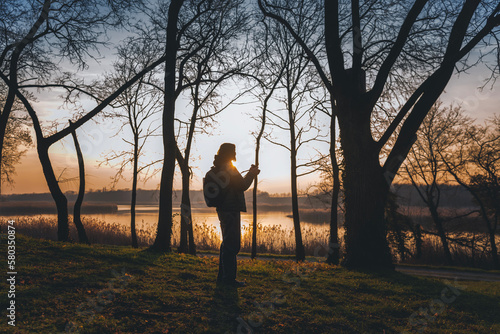 Woman photographer with a camera in her hands and a backpack standing in the spring forest at sunset on the lake © olezzo