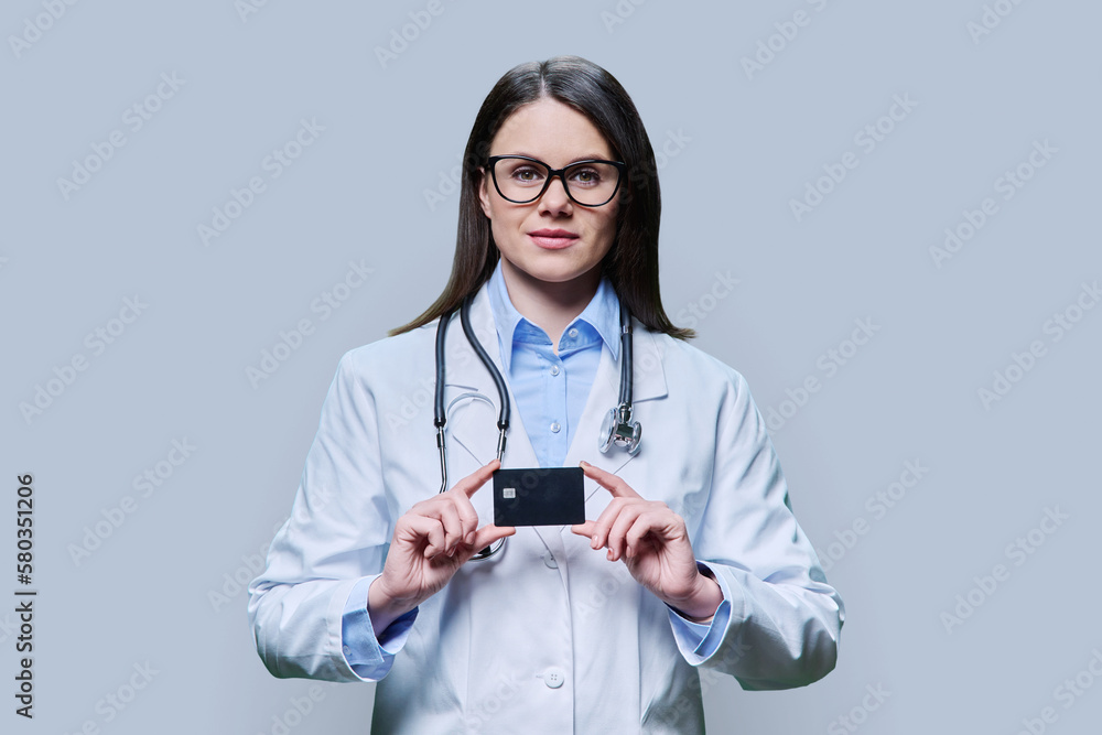 Young woman doctor holding credit card on light gray background Young woman doctor holding credit card on light gray background