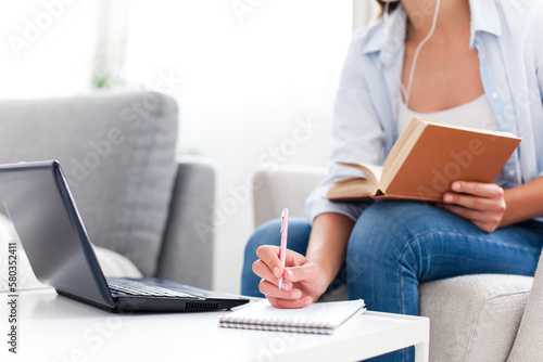 Education online. Student studying from home. Young woman using laptop, earphones, book and Internet. Distance learning, self development in cozy workplace. Freelancer remote working in living room photo