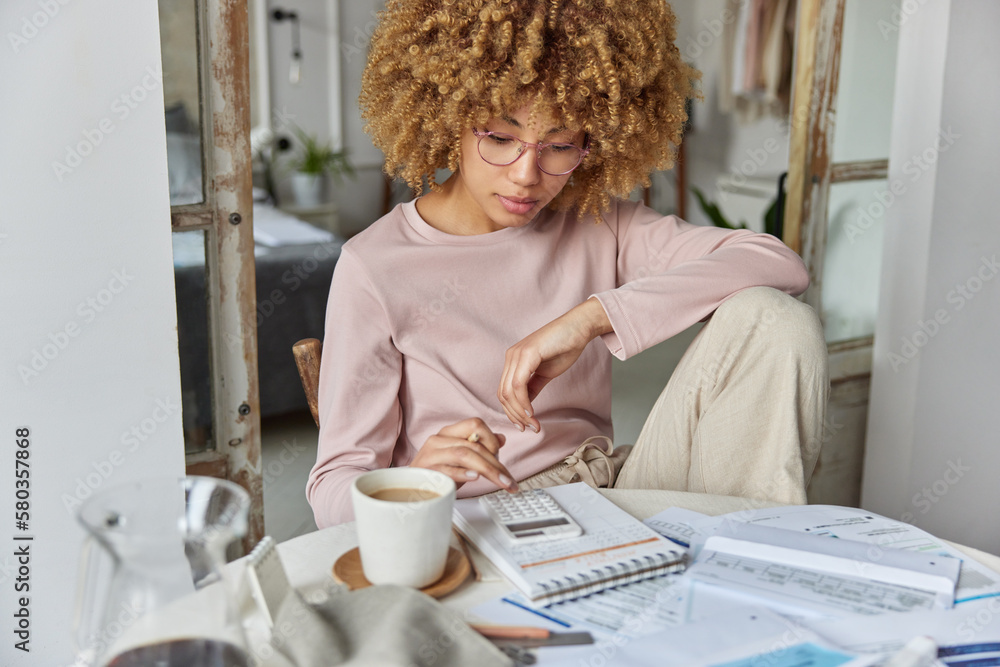 Serious curly haired woman manages household family budget calculates expenditures takes care of finances and savings sits at table with receipts dressed in domestic clothes poses at home office Serious curly haired woman manages household family budget calculates expenditures takes care of finances and savings sits at table with receipts dressed in domestic clothes poses at home office